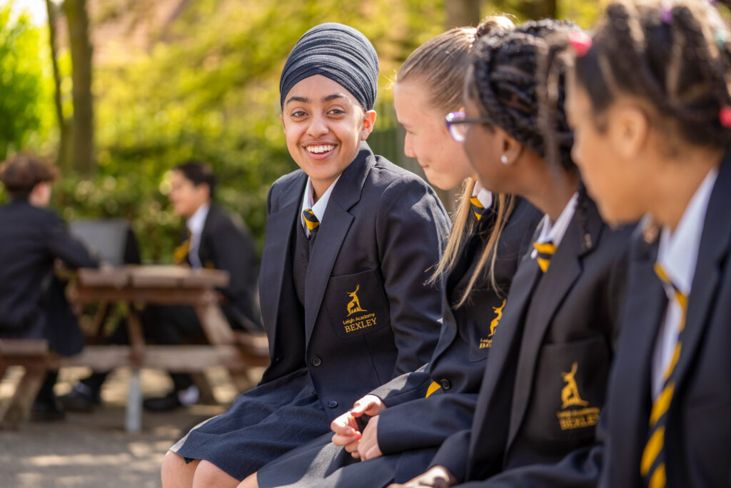 A school student looking at the camera smieling sitting next to 3 friends looking at her. All students are in their school uniform