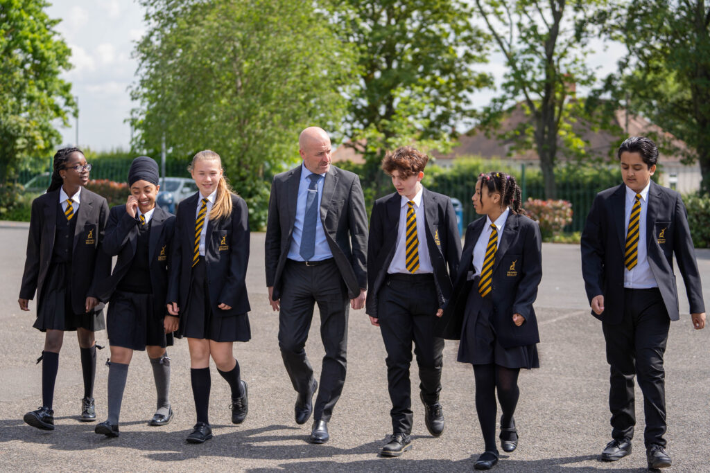 Principal, John Dixon, is seen walking across an outdoor area of the academy grounds alongside six students.