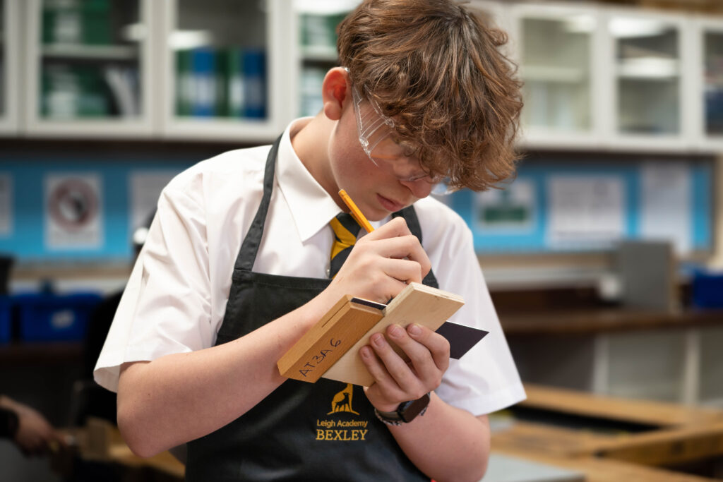 A male student is pictured wearing an apron with the Leigh Academy Bexley logo on, whilst using a pen to mark a piece of wood during a Design & Technology class.