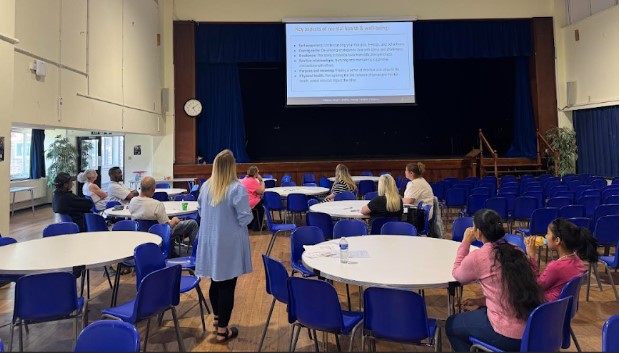 A workshop is taking place in what appears to be a school hall. A person is standing at the front, facing a screen displaying information about "Key aspects of mental health & wellbeing." Attendees are seated at round tables, listening to the presenter.