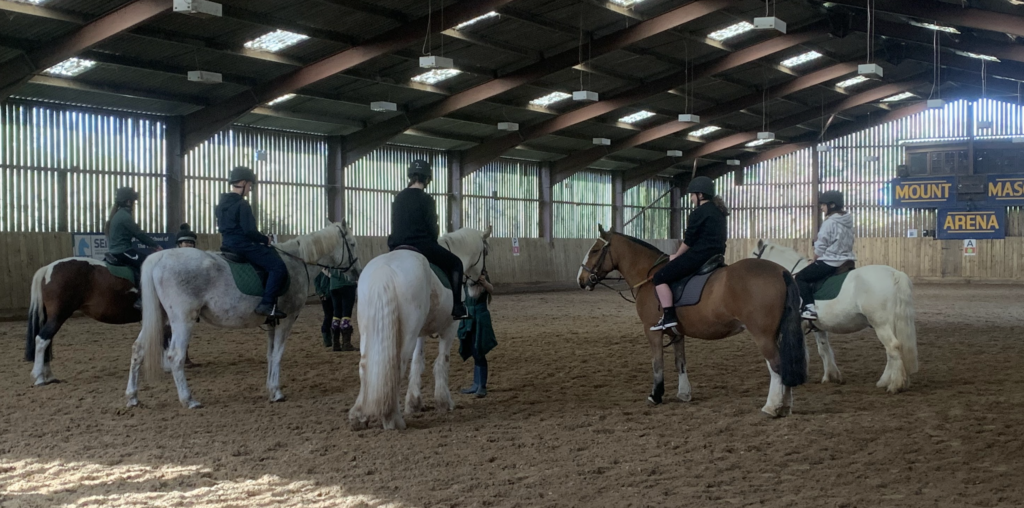 Students stood in a sand school arena on horses