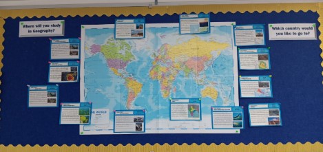 This image displays a blue bulletin board with a gold ruffled border, featuring a large world map in the center. The board seems to be for a geography class, with two questions posed to students: "Where will you study in Geography?" and "Which country would you like to go to?". Around the map are several light blue cards, each highlighting a different location or geographical concept with text and images. While the text on the cards is largely unreadable in detail, they appear to be educational displays related to various parts of the world and geographical features or topics.