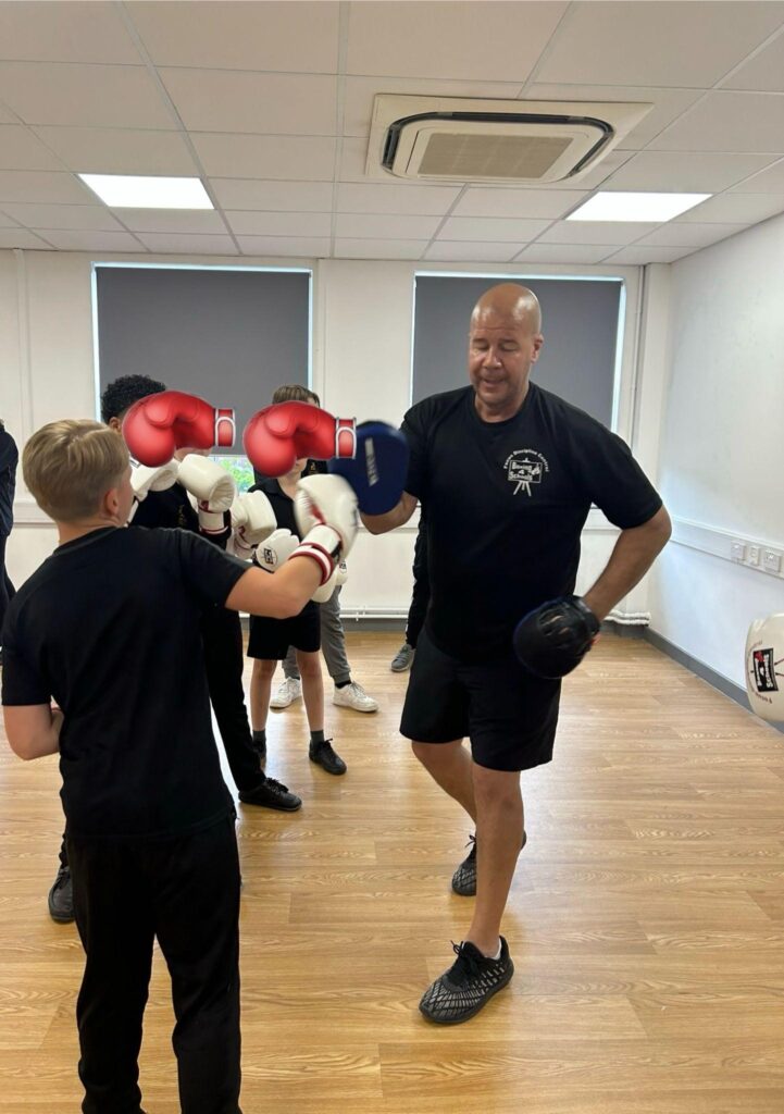 Pupils in PE kits receive boxing lessons from an instructor in a room with wooden flooring