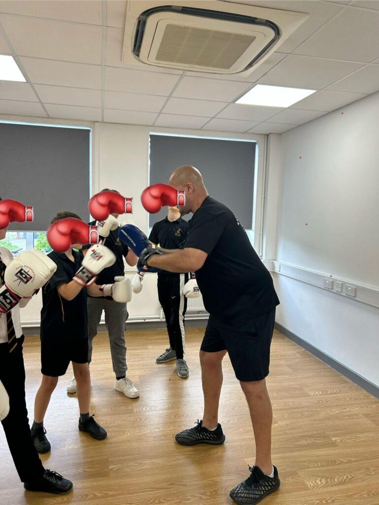 Pupils in PE kits receive boxing lessons from an instructor in a room with wooden flooring