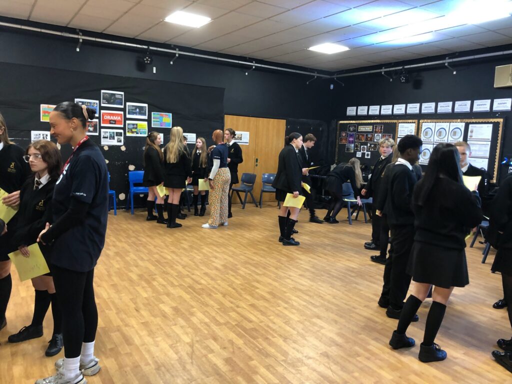 students, most wearing school uniforms (black skirts/trousers and blazers), engaged in what looks like a drama class or rehearsal in a large room with a wooden floor and black walls. BX5.jpg shows a group of students standing, some holding papers, in a busy room. Bx6.jpg focuses on two girls walking across the center of the space, holding yellow sheets of paper, while the rest of the students are seated on the floor around the perimeter, watching. The walls are decorated with posters and signs, some related to "DRAMA."