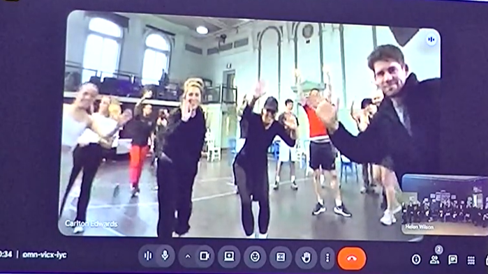 This is a screenshot from a video conference call (Google Meet). The main video feed shows a group of people inside a large room with a high ceiling and wooden floor, possibly a dance or gym studio. They are all participating in a physical activity or dance, with their arms raised in a choreographed move.