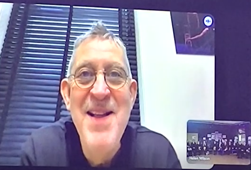 A screenshot from a video conference call. The main feed has shifted to show a close-up of a middle-aged man wearing glasses and a black shirt, smiling.
