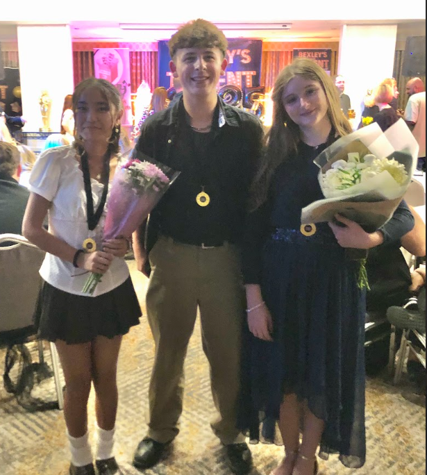These images capture three young people—two girls and a boy—standing together in what appears to be an indoor event space, possibly after a performance or competition. They are all smiling and holding bouquets of flowers and wearing medallions around their necks, suggesting they are winners or performers in the event. The backdrop has a sign that reads "BEXLEY'S TALENT" (partially visible).