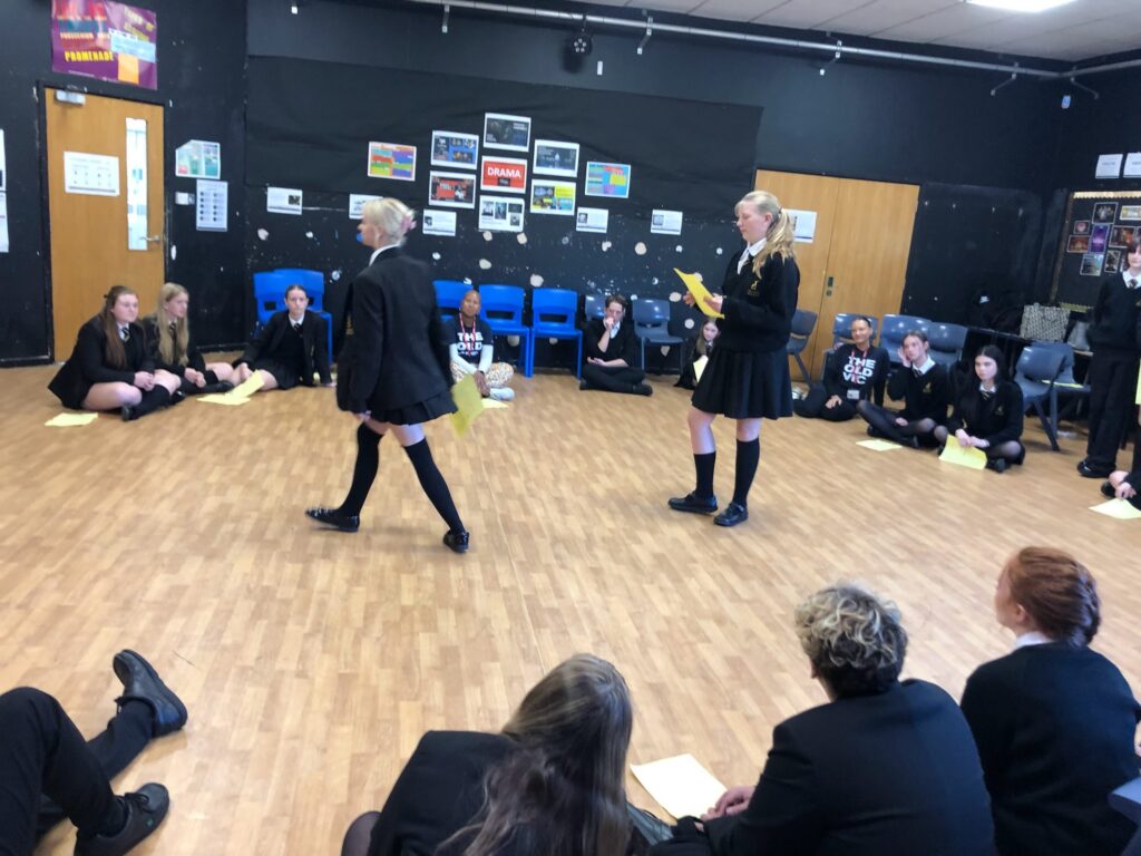 students, most wearing school uniforms (black skirts/trousers and blazers), engaged in what looks like a drama class or rehearsal in a large room with a wooden floor and black walls. BX5.jpg shows a group of students standing, some holding papers, in a busy room. Bx6.jpg focuses on two girls walking across the center of the space, holding yellow sheets of paper, while the rest of the students are seated on the floor around the perimeter, watching. The walls are decorated with posters and signs, some related to "DRAMA."