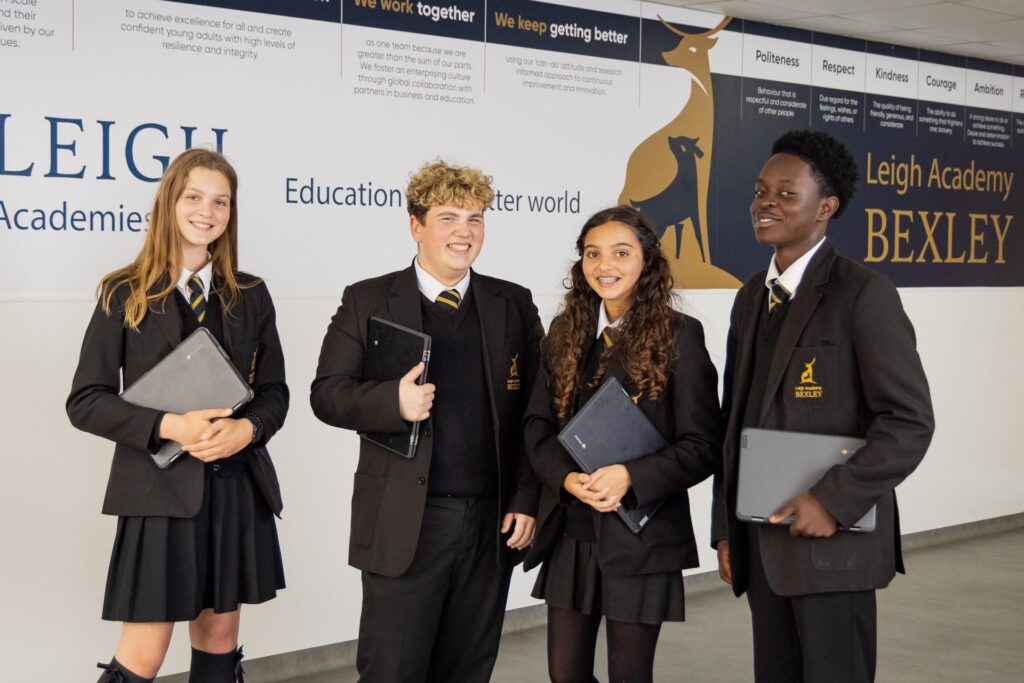 Four students in black and gold school uniforms are standing together, posing for a photo in what appears to be a modern school hallway or entrance. They are all holding electronic tablets or laptops. The background has wall graphics with text about "Leigh Academy Bexley" and school values like Politeness, Respect, Kindness, Courage, and Ambition.