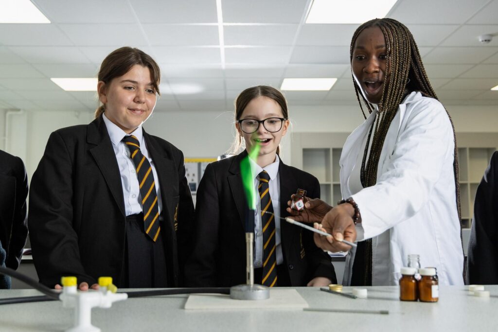 A science laboratory setting where two students in school uniform and a teacher or adult (wearing a white lab coat) are participating in a chemistry experiment. The focus is on a green flame from a Bunsen burner, and the adult is using a wire to hold a chemical into the flame. The students are smiling and observing the process.