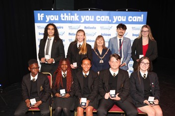 A group of ten individuals is formally posed on a stage with a banner in the background that reads, "If you think you can, you can!" in large white text, alongside smaller logos and words like 'Inspire', 'Motivate', and 'Achieve'. The group includes five young people seated in the front row, wearing dark school uniforms (blazers, white shirts, ties), and five people standing behind them, including one adult female wearing what appears to be a ceremonial chain or badge of office (possibly a mayor or civic leader). The seated students and standing individuals are all holding small, rectangular silver medals or awards. The background is a dark stage area.