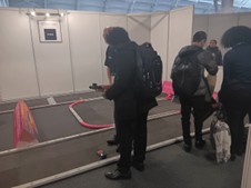 A few young people are gathered around a small, temporary racing track set up in an exhibition hall, possibly for remote-controlled cars or a similar activity. One person is leaning over the track, focusing on a small vehicle, while others stand nearby.