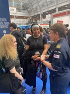 Three people are engaged in a discussion at what appears to be a careers fair or exhibition. A woman in a dark military or uniform shirt, adorned with badges and flags, is talking to two young women in civilian clothes. They are standing on a blue carpet in a large hall with high ceilings. A sign mentioning "RESEARCH ROYAL NAVY CAREERS" is partially visible in the background.