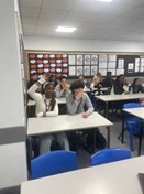 A photo of students seated at desks in a classroom. In the foreground, one student with long hair is looking at the camera, while a student next to them raises their hand. The room is brightly lit, and the walls are decorated with educational posters or visual aids.