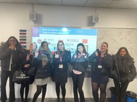 A group of seven young women is standing in front of a projection screen in a modern classroom, all giving a thumbs-up gesture and smiling. The screen displays a title asking, "How to choose a university? Does it matter where?" and features the logos of several universities, suggesting a careers or higher education presentation. Some of the women are wearing dark outdoor coats and carry bags, indicating they may be visitors or about to leave.