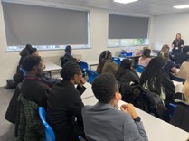 A view from the back of a classroom, showing students seated at desks, listening to a presentation. The heads and shoulders of many students are visible, primarily from the back, as they face the front of the room where a speaker is standing (partially visible in the distance).