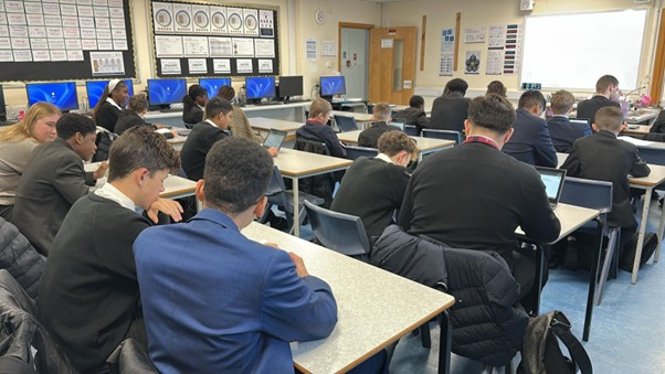 An image taken from the back of a large classroom, showing rows of students, mostly wearing dark school uniforms (blazers and white shirts), seated at desks. Many students have laptops open and are working. The back wall features a line of computer monitors, suggesting this is a computer lab or a technology-equipped room.