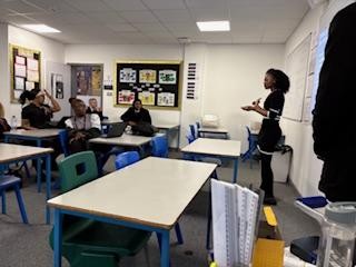 A classroom scene where a young woman is standing near the front of the room, speaking or presenting to a small group of students seated at desks. The photo is taken from a low angle, showing the white desks and blue chairs in the foreground.