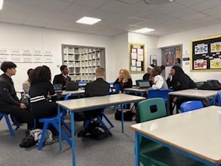 A photo of a modern classroom where students are working in small groups around clustered desks. An adult woman is seated with one group, engaging in discussion. Laptops are visible on the desks, and the room has bright white walls with informational posters and cabinets in the background.