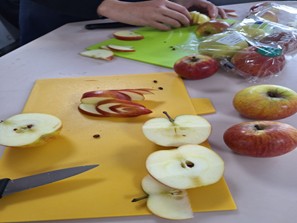 The photo captures a workspace where hands are actively slicing and preparing apples, possibly for the carving activity shown in other images. Several apples, both whole and sliced, are on a light-colored table, alongside bright green and yellow cutting boards. Halved apples and thin, curved slices are visible on the boards, with a knife resting on the yellow board.