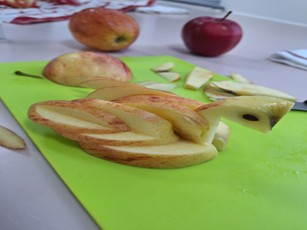 A close-up of a fruit carving in progress on a bright lime-green cutting board. The carving, made from pale apple slices with red skin, is shaped like a bird or swan, with a head carved out using an apple seed for the eye. Whole and cut apple pieces are visible in the background.