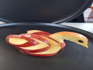 A close-up view of an intricate food carving made from slices of a red apple. The slices are layered and curved to resemble a swan or bird, with one apple seed used to represent the eye. The carving rests on a dark, circular surface, possibly a plate or platter.