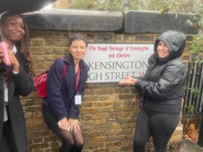 Three individuals are standing outside under an overhang or canopy (suggesting light rain), next to a large, weathered brick wall sign. The sign, which is partially obscured, reads: "The Royal Borough of Kensington and Chelsea" and "KENSINGTON HIGH STREET W8". They are smiling, and one person on the right is pointing toward the sign.