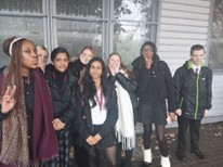 A group of seven young people, standing outside against a backdrop of glass or metal-framed windows, are posing for a picture. They are mostly wearing dark coats, jackets, and scarves. Some are smiling or making playful gestures, such as one young woman holding up a peace sign with her fingers.