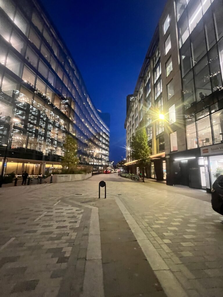 A modern city street scene at night between two large, curved office buildings with glass facades. The buildings are brightly lit inside, and the street below is illuminated by streetlights.