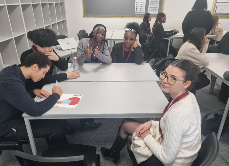 A small group of students and an adult facilitator are working together at a table. One student is writing on a paper featuring a diagram, while others are engaging with the activity and posing for the camera.