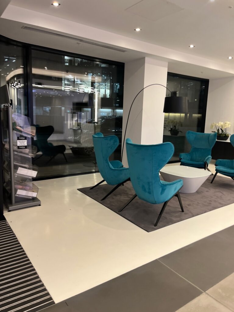 An image of a modern, minimalist office or lobby interior. The space features a seating area with distinctive turquoise-blue velvet wingback chairs, a white circular table, and a large black arc floor lamp, with large windows overlooking the exterior.