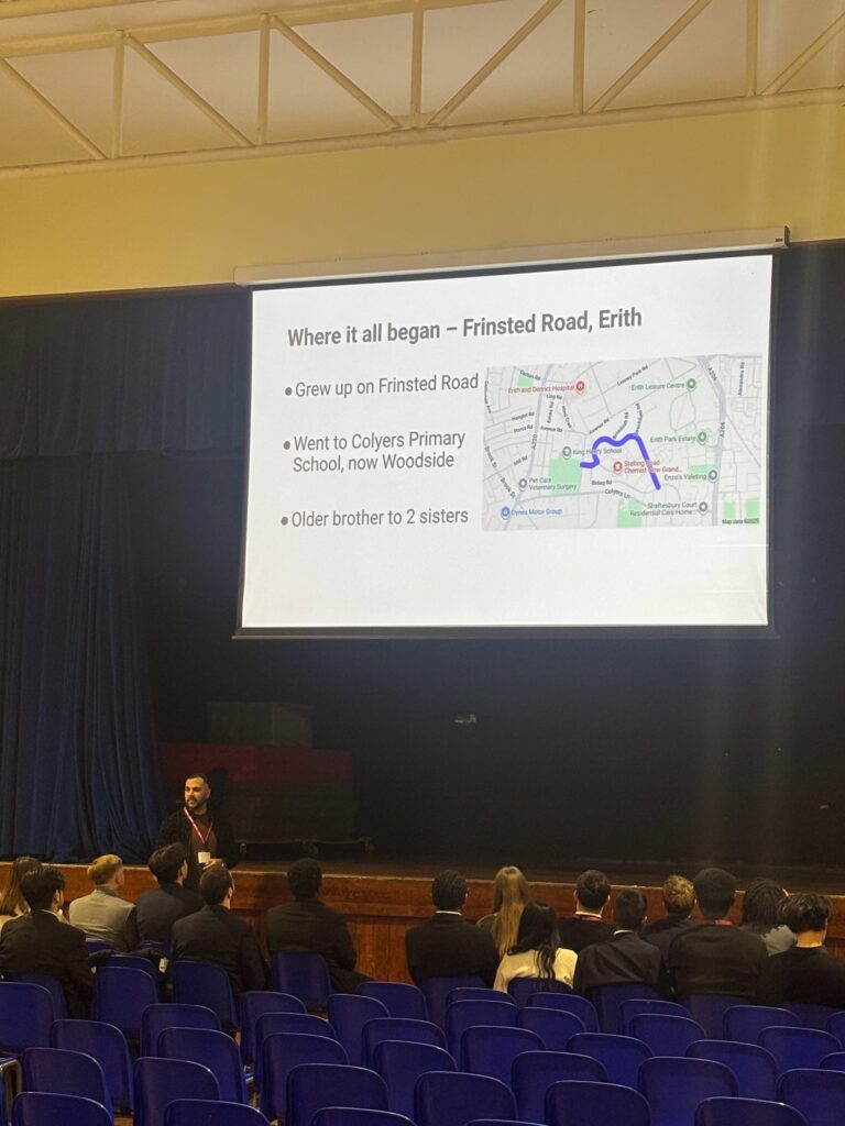 A speaker is giving a presentation on a stage in a large hall to an audience of students, who are dressed in school uniforms. The presentation slide is titled "Where it all began – Frinsted Road, Erith," detailing aspects of the speaker's background.