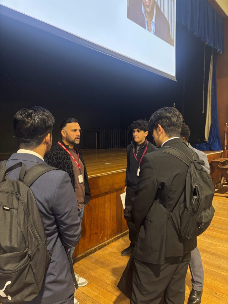 A man is engaging with a small group of students on the stage floor after the presentation. The students, wearing blazers and carrying backpacks, are clustered around the speaker, likely asking questions or networking.
