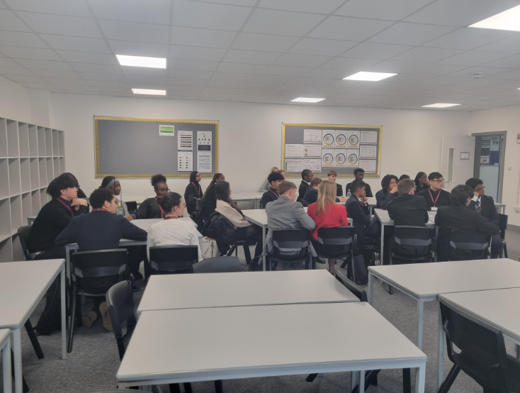 A classroom is full of students and a few adults (some in business attire) seated in small groups at tables, facing forward. The overall setting appears to be the beginning of a presentation or a joint activity in a modern, well-lit teaching space.