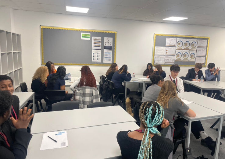 Students are seated in small groups facing each other across tables, engaging in discussion. The layout suggests a collaborative and interactive workshop environment where they are talking through a task or topic.
