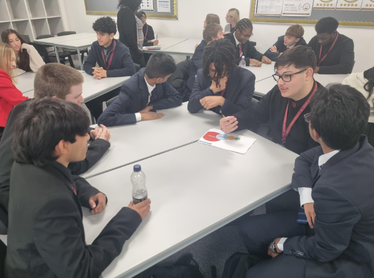 A group of male students, dressed in school blazers, is deeply engaged in a discussion around a circular diagram on their table. They are leaning in, showing focused engagement with the group task.