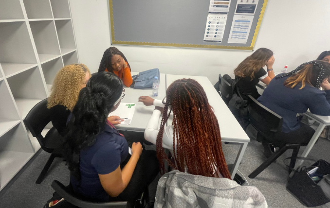 A group of female students is sitting closely together and working on a task at a table. They are focused on materials on the table and interacting with each other, reinforcing the collaborative nature of the session.