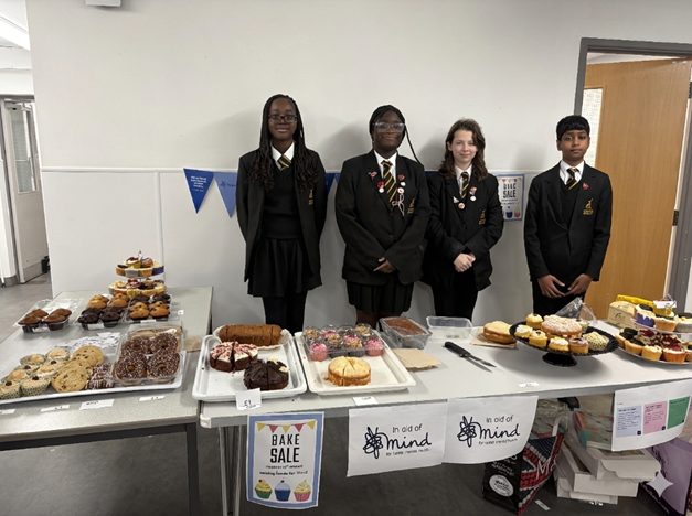 Four students in school uniforms stand behind a table laden with cakes, muffins, and cookies at a bake sale fundraiser in aid of the charity Mind.