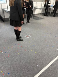 Students in uniform observe an object on a carpet with colored dots.