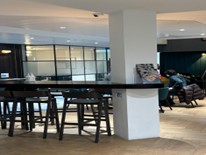 An interior shot of a modern common area or cafeteria featuring a high wooden bar table with stools and a large white structural pillar.