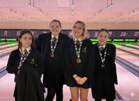 Four female students in school uniforms stand together in a bowling alley wearing medals around their necks.