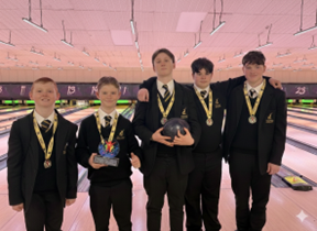 Five male students in school uniforms pose in a bowling alley; they are wearing medals, and one student holds a bowling ball while another holds a trophy.