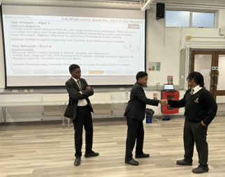 Three students in school uniforms participate in a drama activity in front of a projector screen showing text related to Shakespeare's The Tempest. Two students are shaking hands while a third watches with arms crossed.