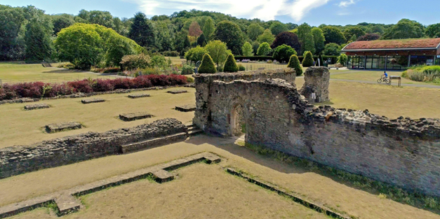 An elevated landscape view of stone ruins, possibly an old abbey or fortification, situated in a large grassy park area with trees and a modern building in the background.
