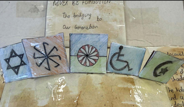 A close-up of the central part of the Holocaust memorial display, showing five hand-drawn squares with symbols including the Star of David, a Romani wheel, a wheelchair symbol, and a crescent and star.