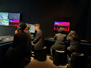 Students in school uniforms sitting at a long desk in a dark room, working on computers and viewing multiple screens displaying video content.