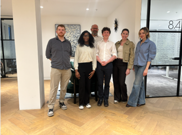 A group of six people (four adults and two teenagers) standing together and posing for a photo in a modern, brightly lit office or school hallway.