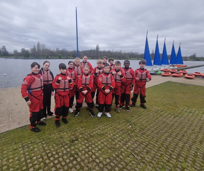 A group of students wearing red drysuits and pink life jackets stands on a grassy bank near a lake with sailboats in the background.