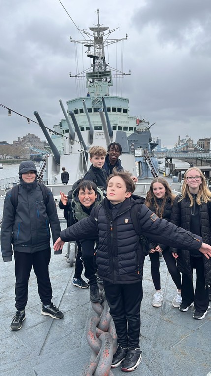 A group of students poses on the deck of a large grey naval ship, with the ship's bridge and several large gun turrets visible behind them.